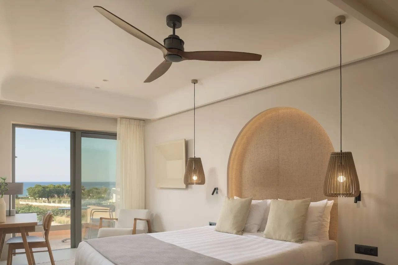 Schlafbereich mit Deckenventilator aus Holz und Balkontür mit Blick auf das Meer.