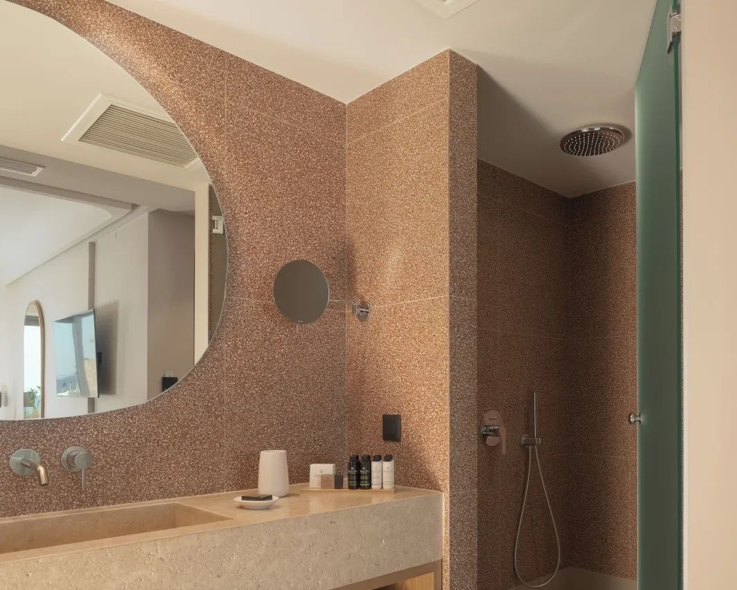 Badezimmer mit beige gefliesten Wänden, rundem Spiegel, Waschtisch und Regendusche.