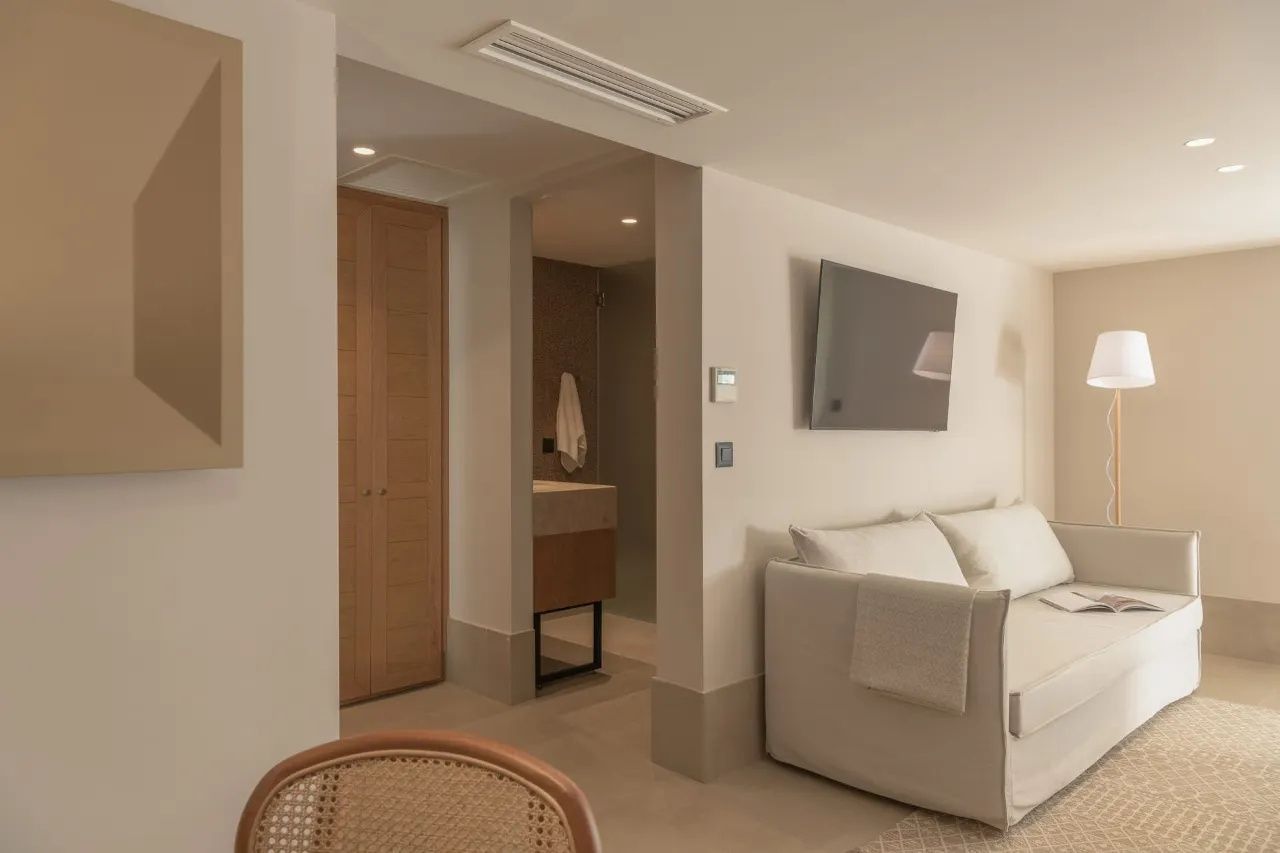 Modern eingerichteter Wohnbereich einer Suite mit hellem Sofa, Wandfernseher und Blick zum Badezimmer.