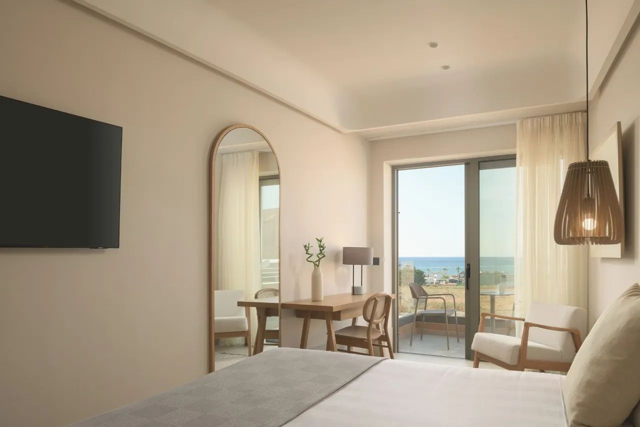 Zimmer mit Standspiegel, Holzschreibtisch und großer Glasbalkontür mit Meerblick.