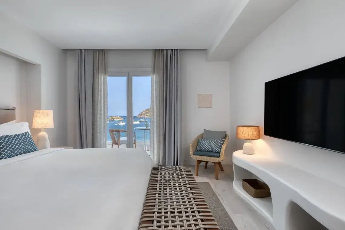 Heller Schlafraum mit Doppelbett, Sitzbank und Blick durch die Balkontür auf das Meer.