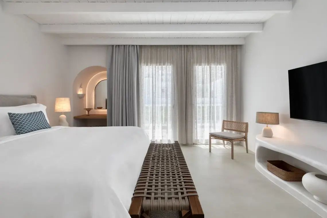 Helles Hotelzimmer mit weißem Doppelbett, geflochtener Sitzbank und hellen Vorhängen vor der Balkontür.