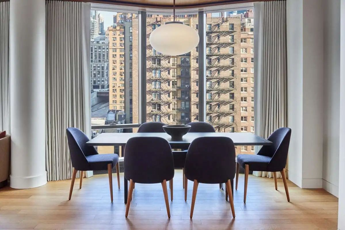 Essbereich mit großem Tisch, dunklen Stühlen und großen Fensterfronten mit Blick auf Stadtgebäude.