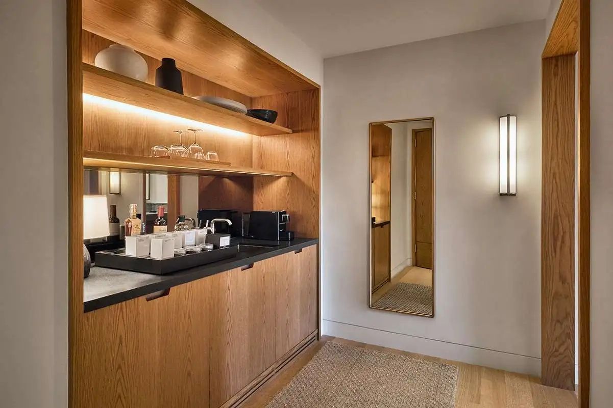 Holzverkleidete Kitchenette mit beleuchteten Regalen, Getränkeauswahl und einem Ganzkörperspiegel.