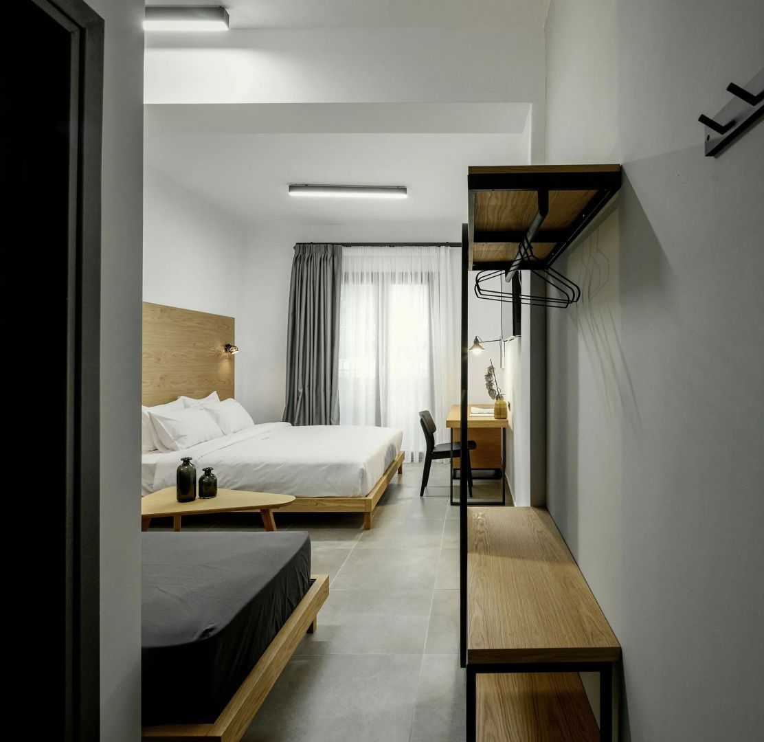 Offenes Hotelzimmer mit Doppelbett, weiterem Bett und schwarzem Metallregal im Vordergrund.