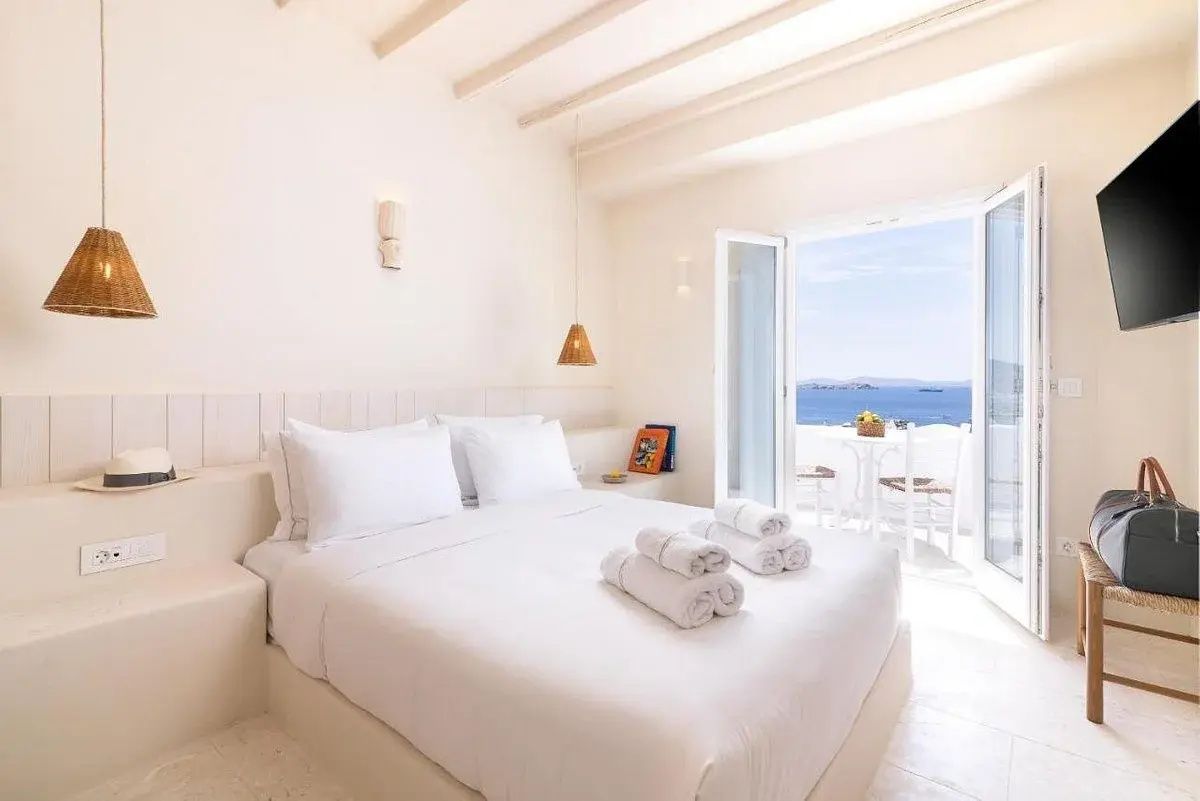 Helles Hotelzimmer in Weiß mit Doppelbett und Blick durch die Balkontür auf das Meer.