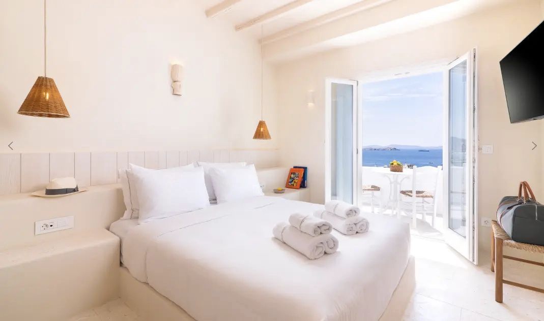 Helles Hotelzimmer in Weiß mit Doppelbett und Blick durch die Balkontür auf das blaue Meer.