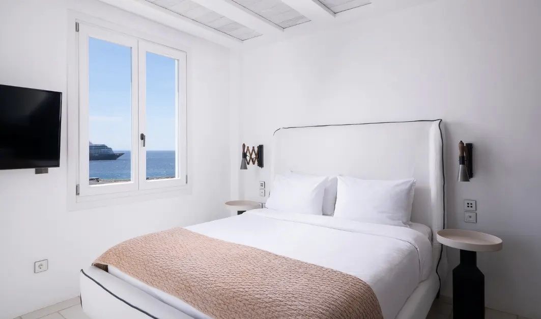 Weiß eingerichtetes Doppelzimmer mit Blick durch das Fenster auf das blaue Meer.
