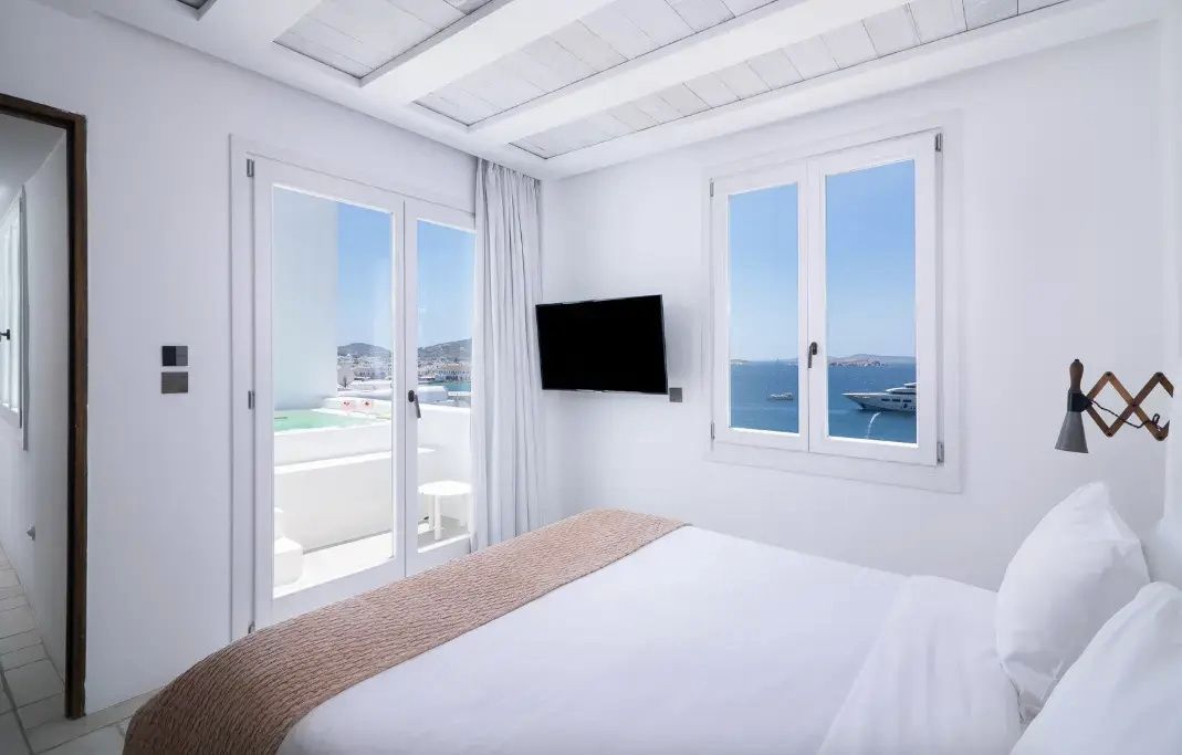 Helles Schlafzimmer mit Holzdecke, Doppelbett, Fernseher und Meerblick durch Fenster und Terrassentür.