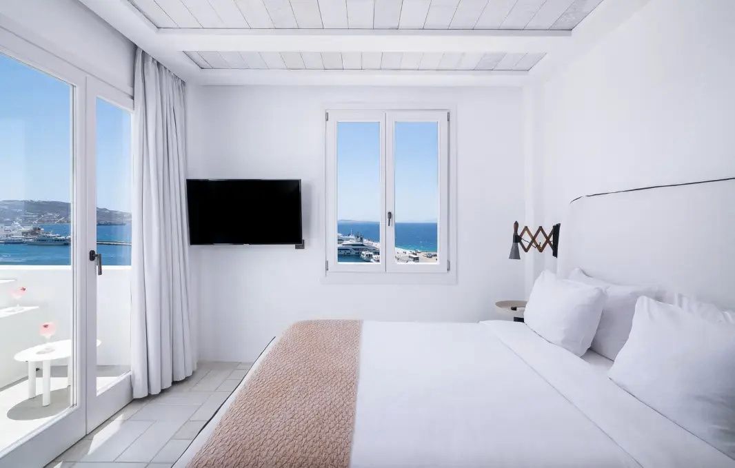 Weißes Hotelzimmer mit Doppelbett, Flachbild-TV an der Wand und Fenster mit Blick auf das Meer.