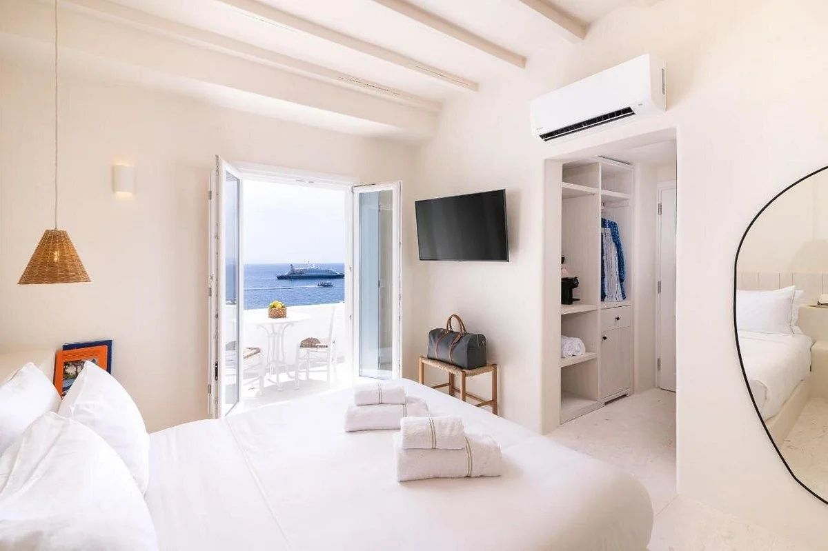 Weiß eingerichtetes Doppelzimmer mit Fernseher, offenem Schrank und Meerblick vom Balkon.