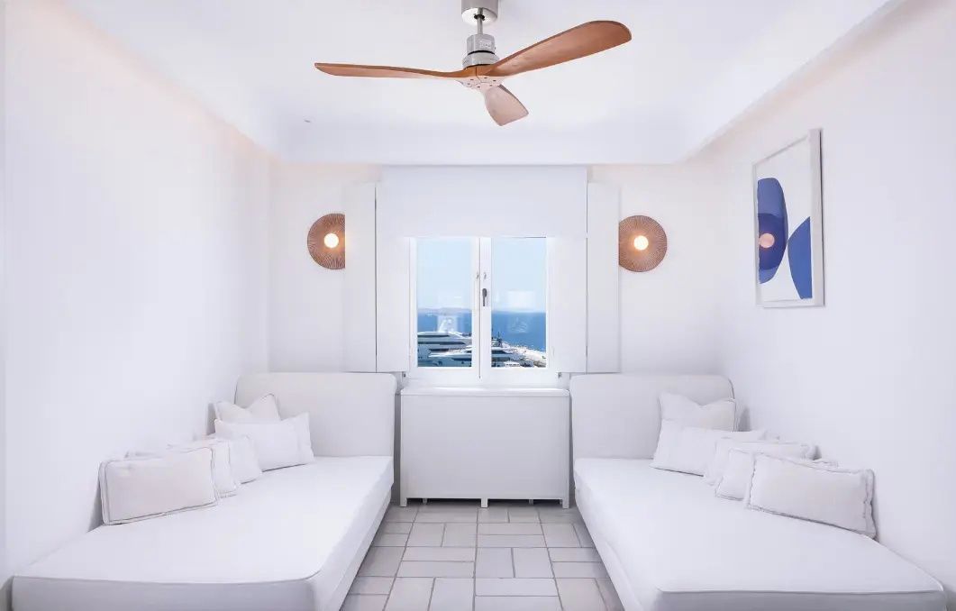Wohnbereich mit zwei weißen Sofas, Deckenventilator aus Holz und Fenster mit Aussicht auf das Meer.