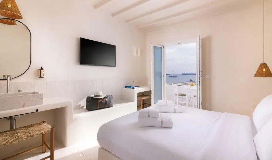 Weißes Zimmer mit Fernseher an der Wand, Sitzbank und Balkontür mit Blick auf das Meer.