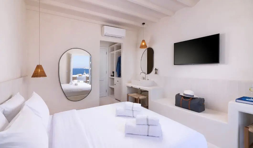 Weißes Hotelzimmer mit Doppelbett, offenem Schrank, Waschtisch und geformtem Spiegel.