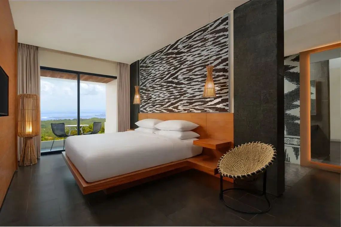 Doppelbett im Hotelzimmer mit gemusterter Kopfteilwand, Flechtsessel und Fenstertür zum Balkon.