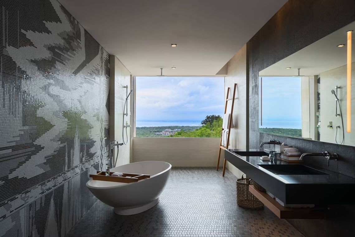 Badezimmer mit ovaler Badewanne, breitem Waschtisch und großem Fenster mit Blick aufs Meer.