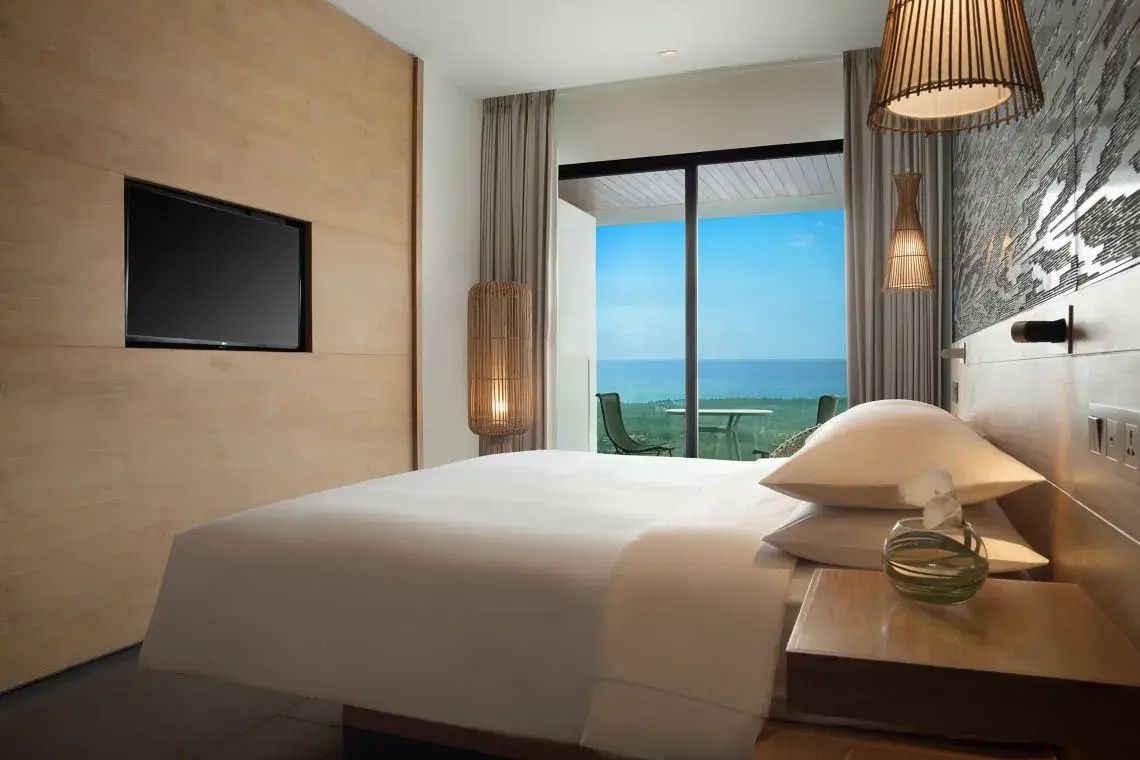 Doppelbett in hell eingerichtetem Hotelzimmer mit Wandfernseher und Aussicht auf das Meer.