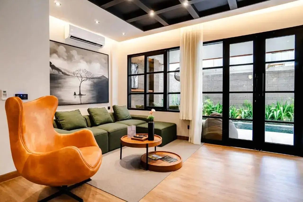 Wohnbereich mit grünem Sofa, orangefarbenem Sessel und großen Fensterfronten.
