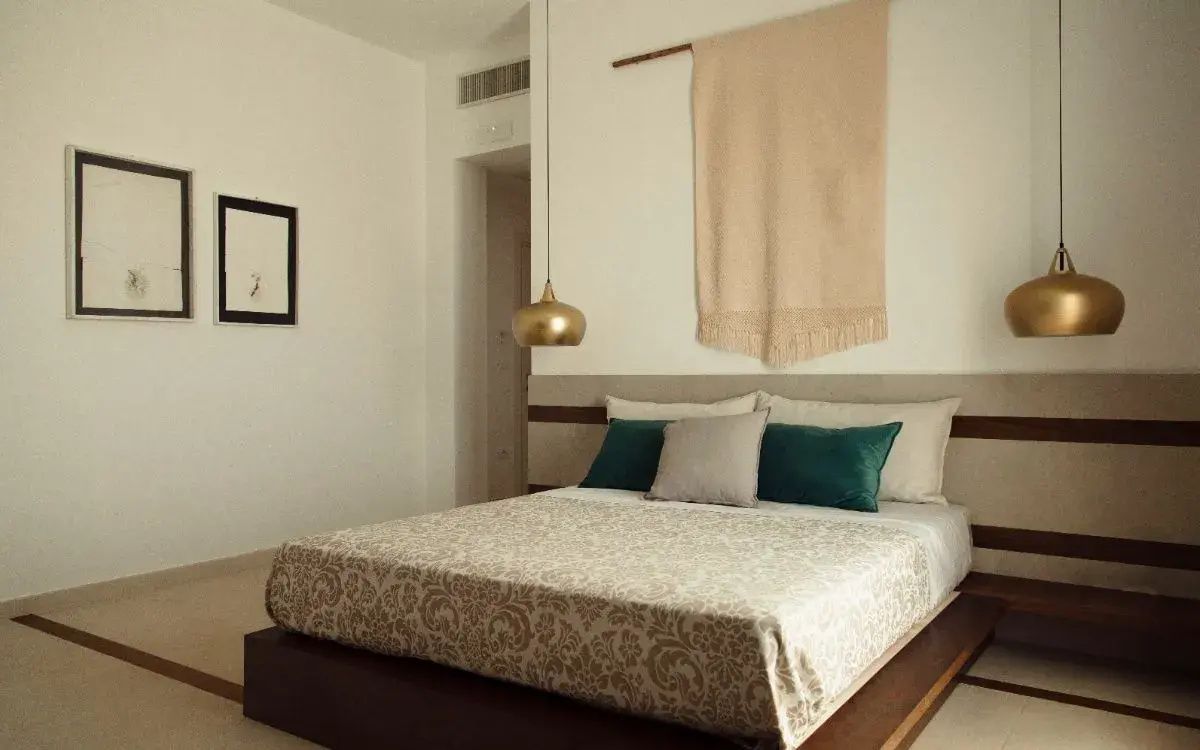 Doppelbett mit gemusterter Tagesdecke, grünen Kissen und Wandbehang in einem hellen Hotelzimmer.