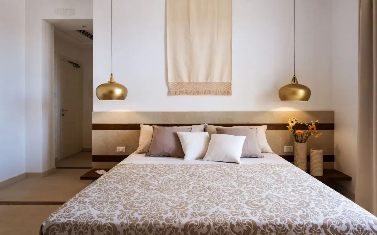 Doppelbett mit gemusterter Tagesdecke, Kissen und goldenen Hängeleuchten im Hotelzimmer.