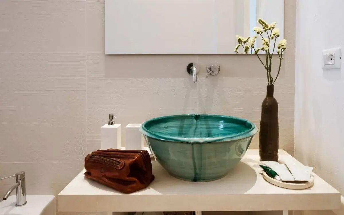 Türkisfarbenes Waschbecken aus Keramik auf einer hellen Ablage mit Kosmetiktasche und Blume.