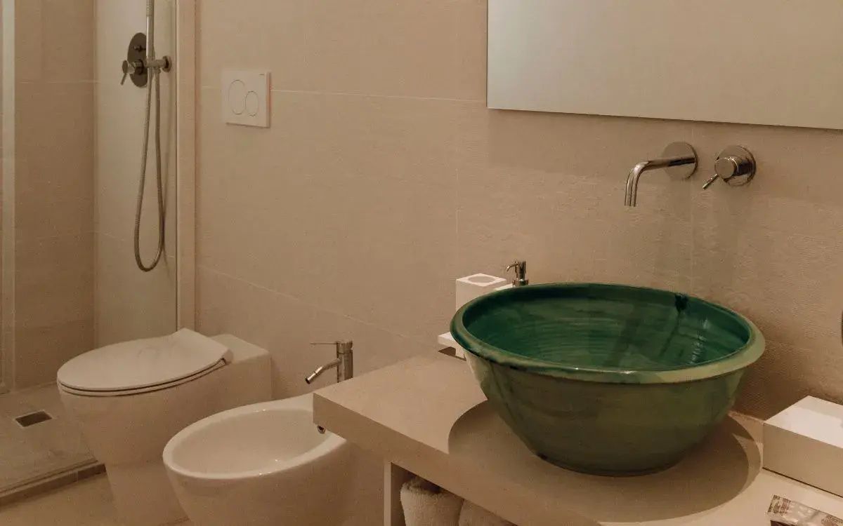 Badezimmer mit grünem Aufsatzwaschbecken, Bidet und hell gefliesten Wänden.