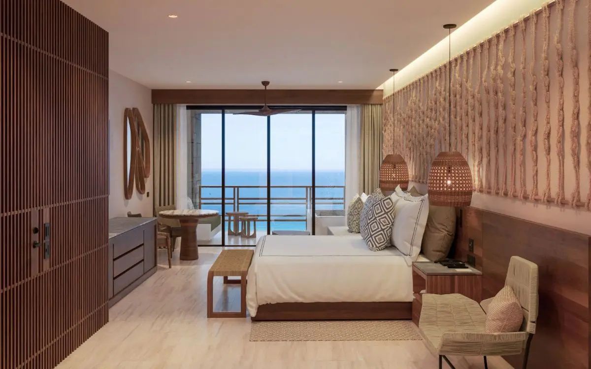 Helles Hotelzimmer mit Doppelbett, Flechtlampen und Glasfront mit Blick auf das Meer.