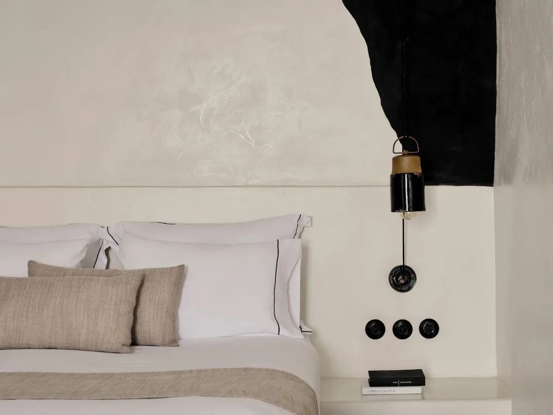Doppelbett mit beige-weißen Kissen und schwarzer Leuchte an der weiß verputzten Wand.