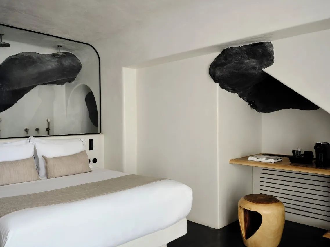 Doppelbett in modernem weißem Hotelzimmer mit dunklen Felsakzenten und Holzschemel.
