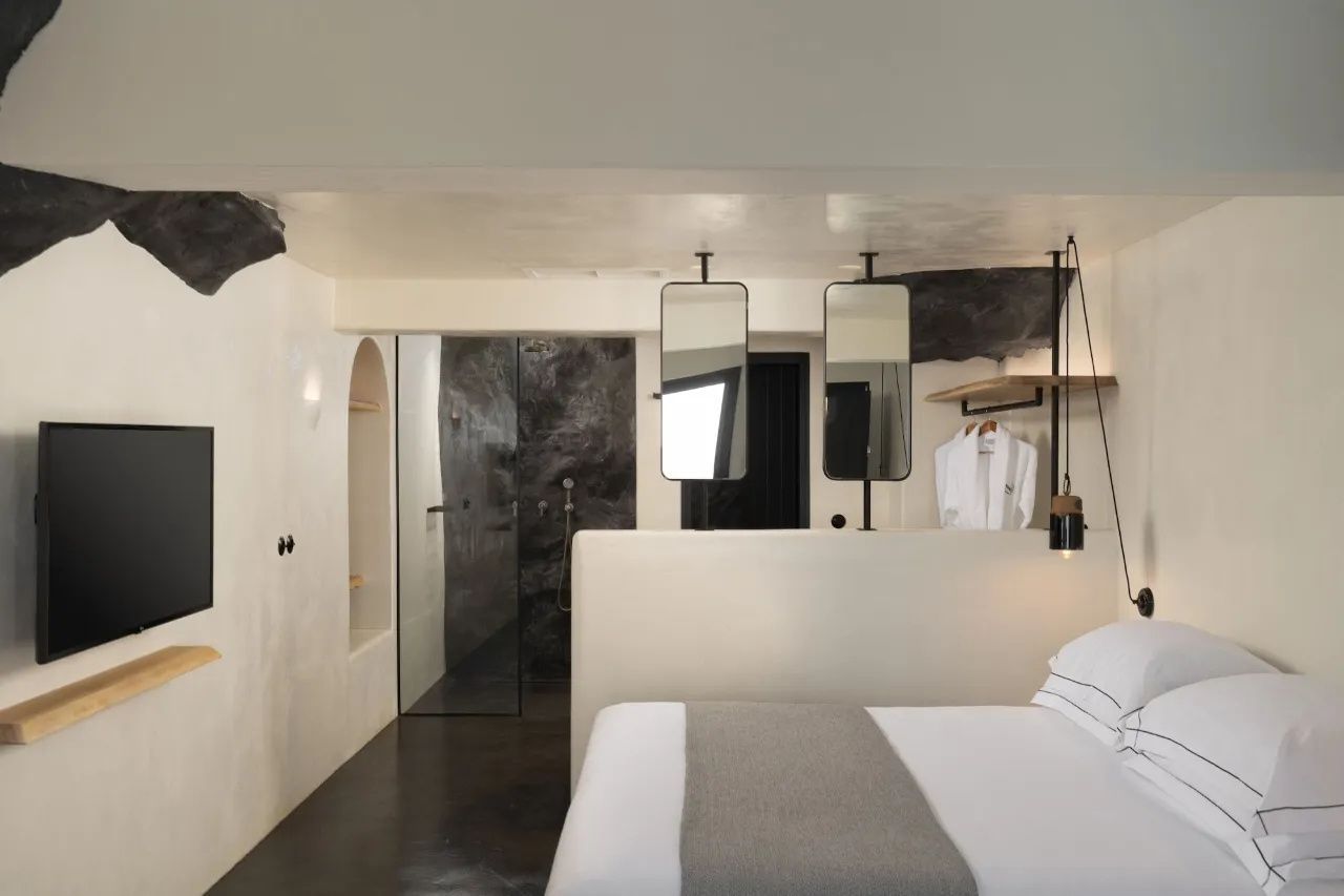Hotelzimmer mit weißem Bett, offener Dusche mit Natursteinwand und zwei hängenden Spiegeln.