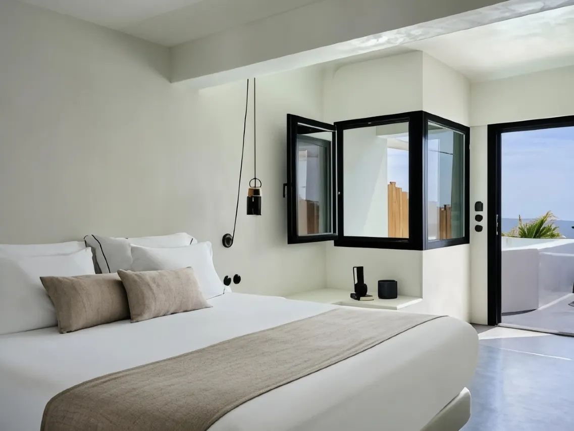 Doppelbett mit hellbrauner Tagesdecke und Kissen vor weißer Wand und geöffnetem Fenster.