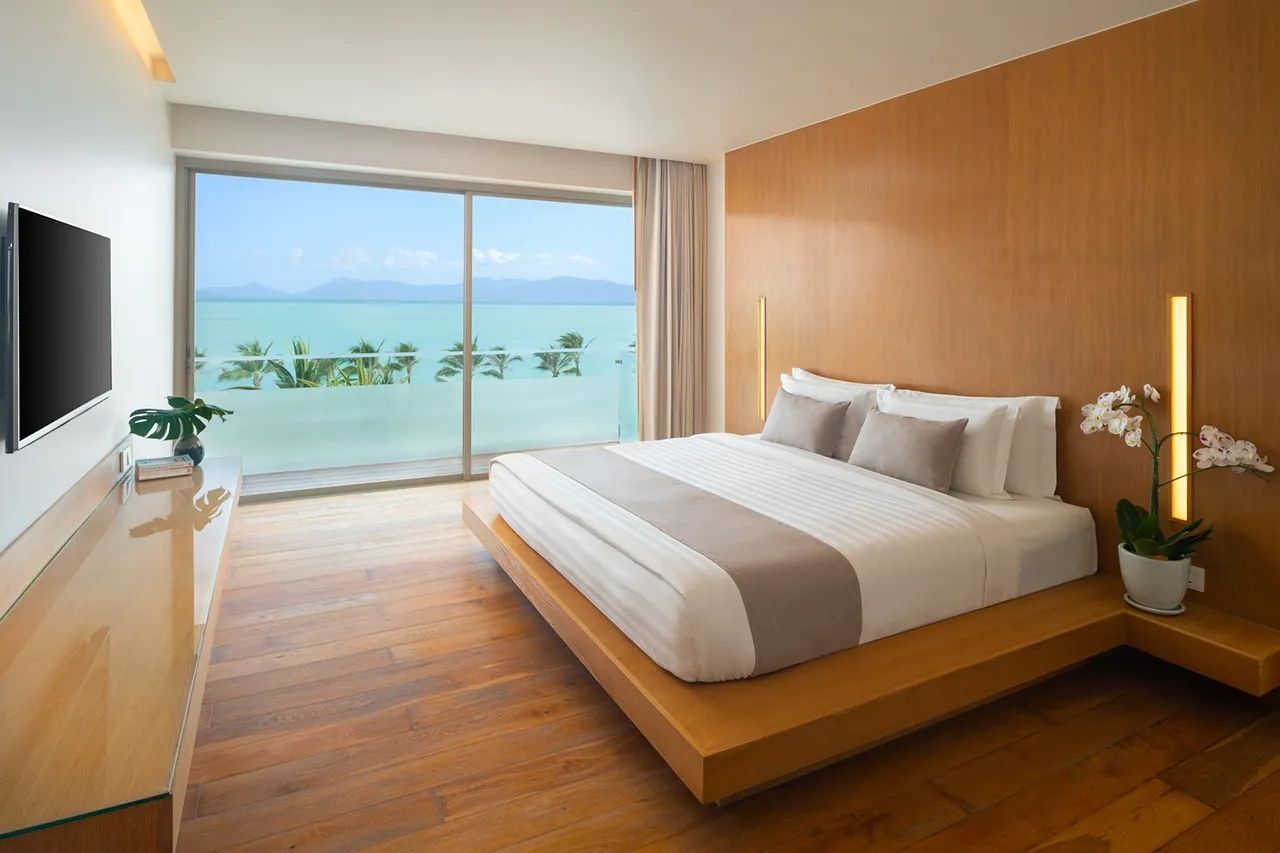 Doppelbett aus Holz mit weißer Bettwäsche und freiem Blick durch das Glasfenster auf das Meer.