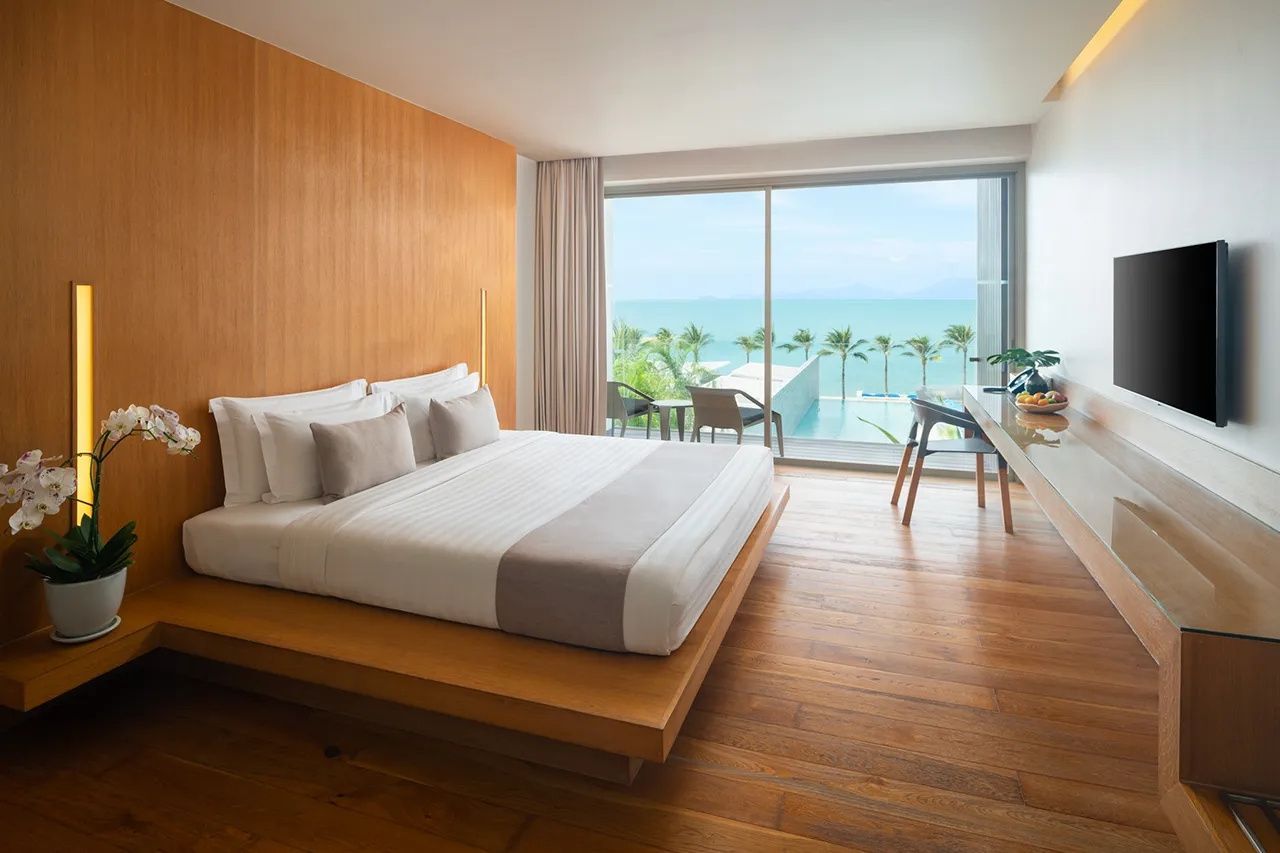Helles Hotelzimmer mit Doppelbett, Holzboden und Balkon mit Blick auf das Meer und den Pool.