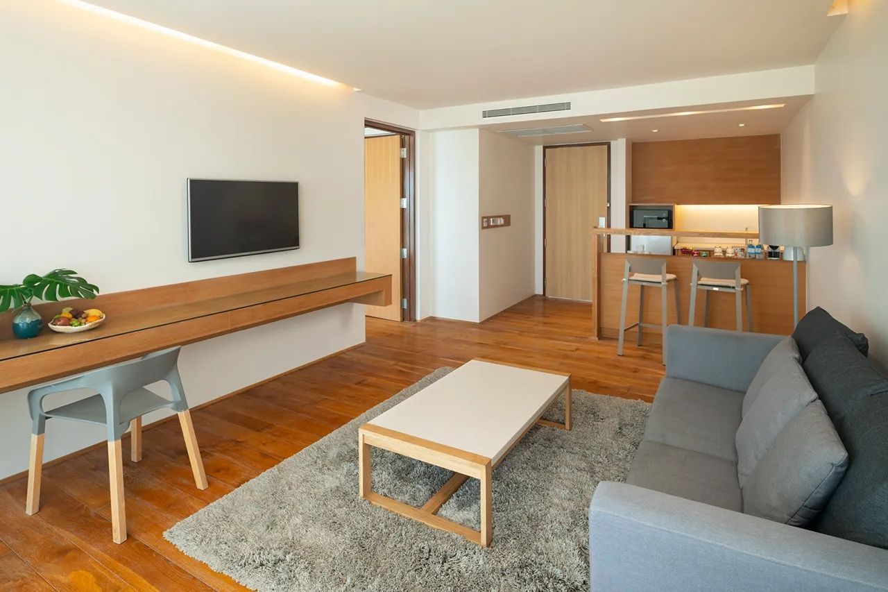 Wohnbereich mit grauer Couch, Tisch auf Teppich, Wandfernseher und kleiner Küchenzeile dahinter.