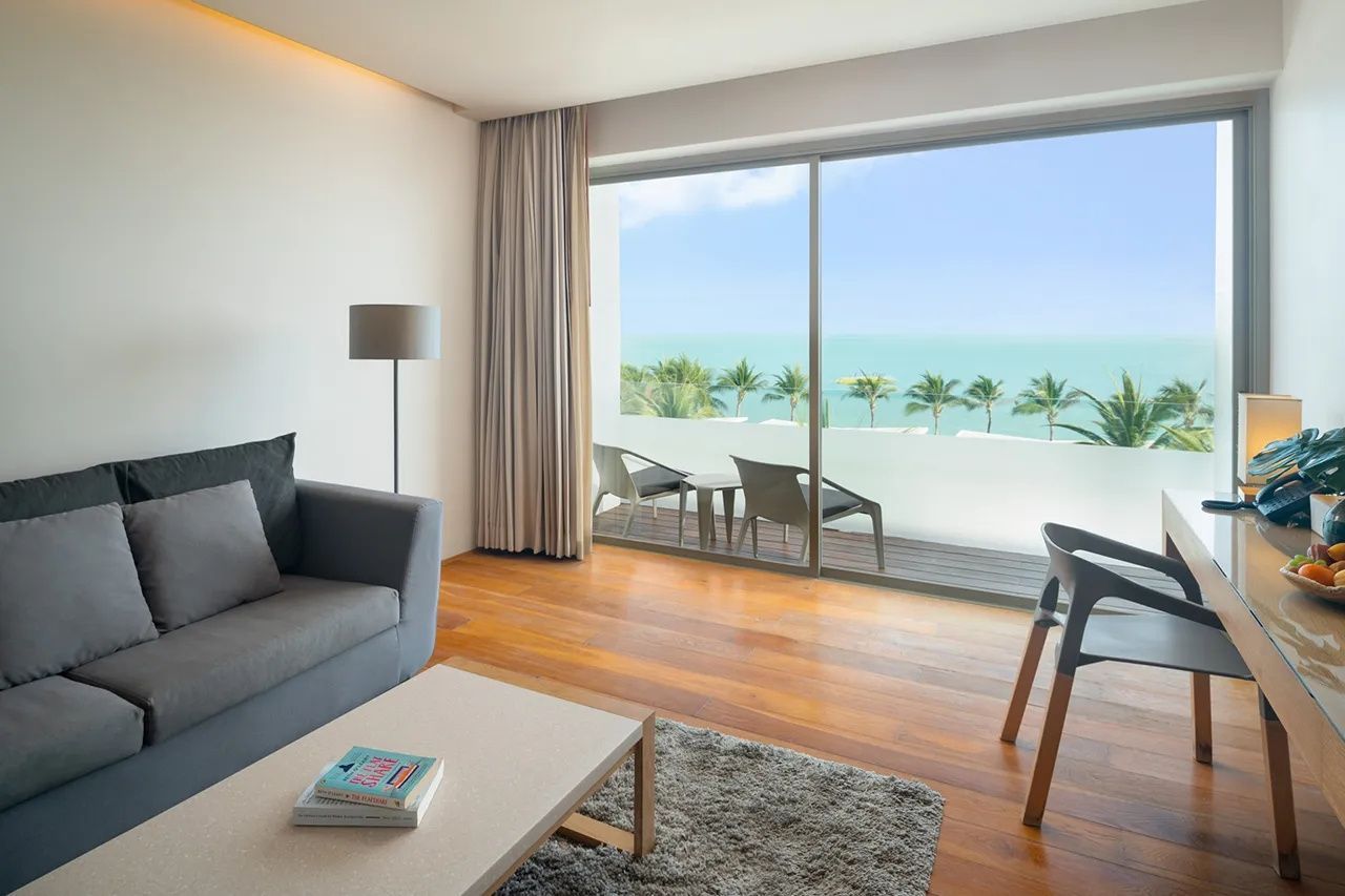 Blick aus dem Wohnbereich über den Balkon mit zwei Stühlen direkt auf Palmen und das blaue Meer.