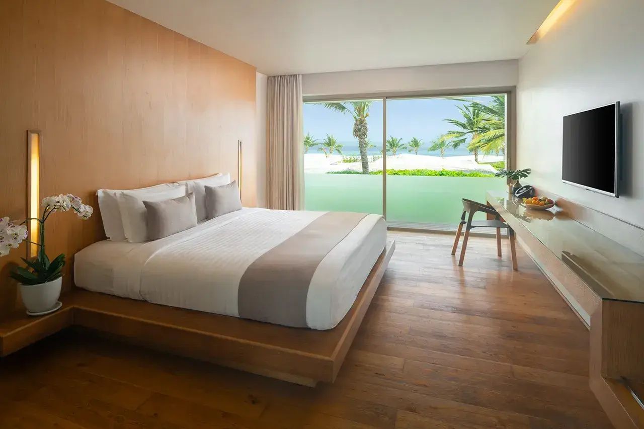 Schlafbereich eines Hotelzimmers mit Holzfussboden und Ausblick auf Strand und Palmen.