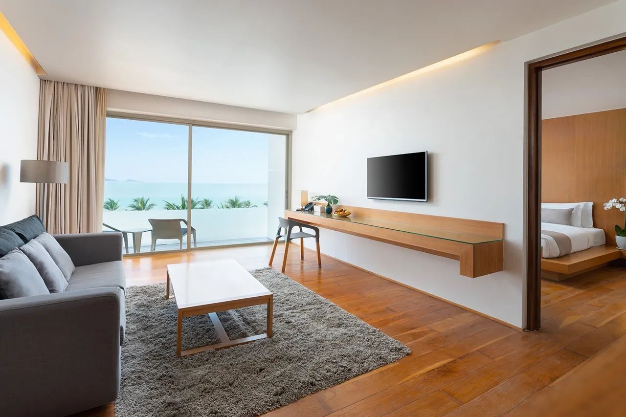 Hell ausgeleuchteter Wohnraum mit hölzernem Schreibtisch, Fernseher und Meerblick durch die Schiebetür.