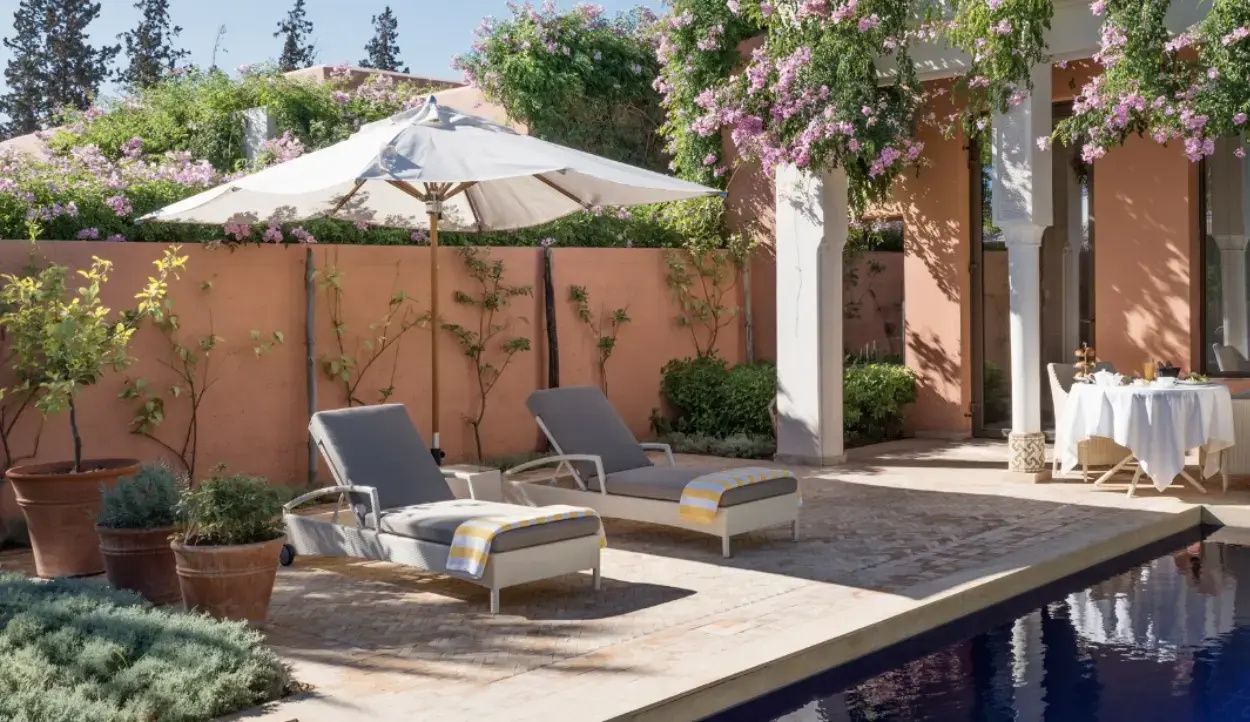 Zwei Sonnenliegen mit Sonnenschirm an einem Pool vor einer rosa Mauer mit Pflanzen.