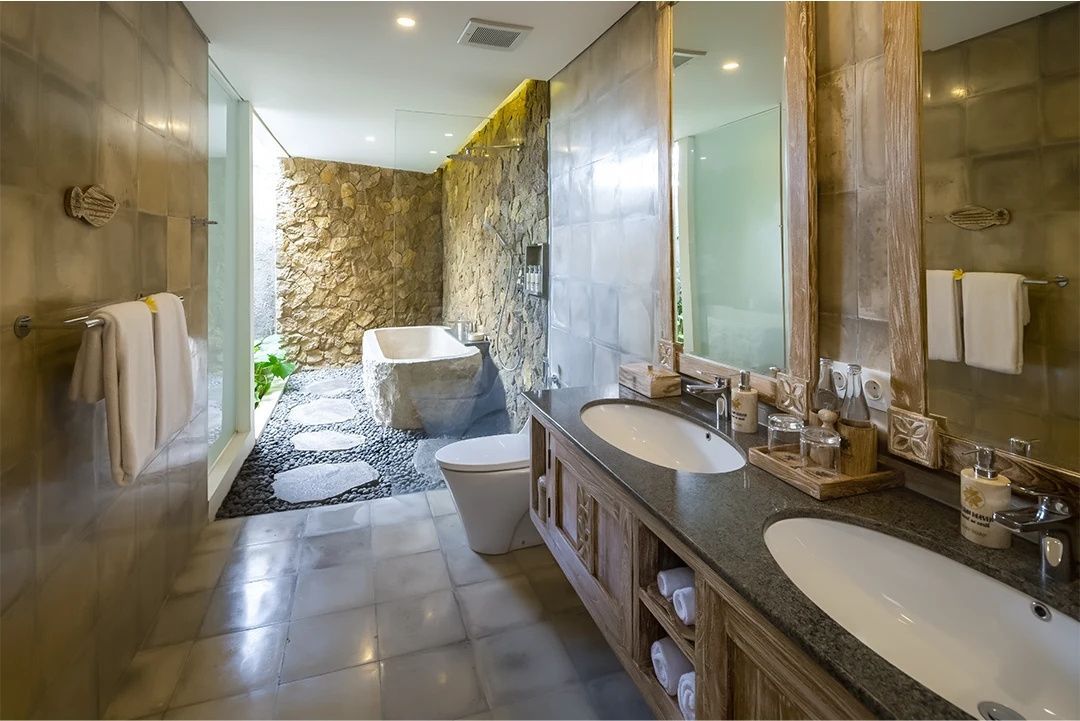 Badezimmer mit doppeltem Waschtisch, Toilette und freistehender Badewanne vor einer Steinwand