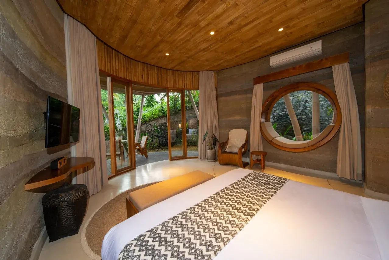 Schlafzimmer mit Holzdecke, Doppelbett, rundem Fenster und großen Glas-Schiebetüren zur Terrasse.