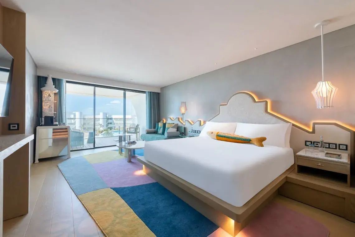 Modernes Hotelzimmer mit großem Doppelbett, buntem Teppich und Glasfront zum Balkon.