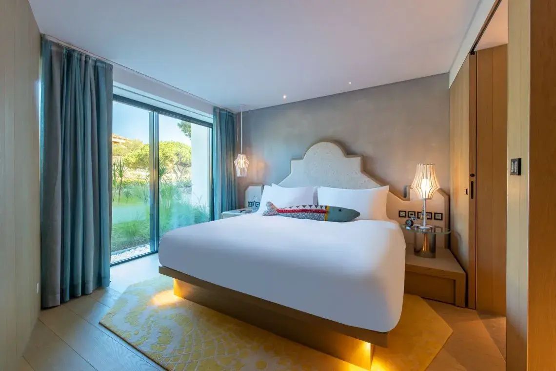 Hell beleuchtetes Hotelzimmer mit einem Doppelbett und bodentiefen Fenstern zum Garten.