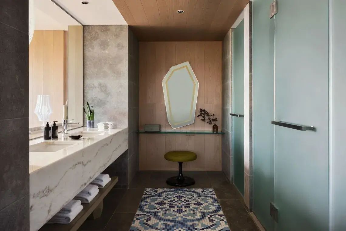 Badezimmer mit Marmorwaschtisch, spitzem Wandspiegel, Hocker und gemustertem Teppich.