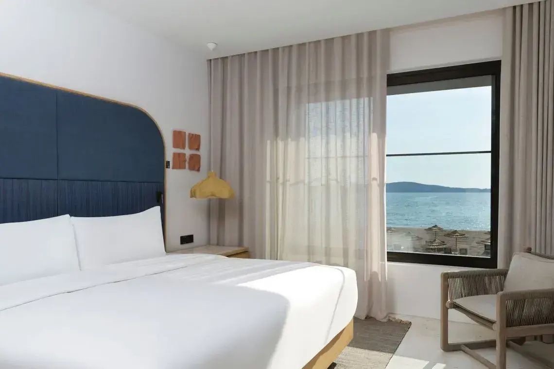 Helles Hotelzimmer mit Doppelbett, blauem Kopfteil und Blick durch das Fenster auf das Meer.