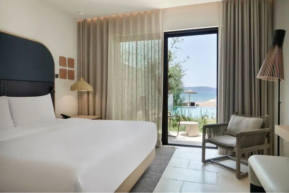 Hell eingerichtetes Hotelzimmer mit Doppelbett und Blick auf Terrasse, Pool und Meer.