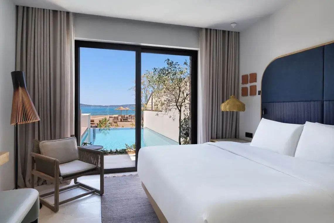 Helles Hotelzimmer mit Doppelbett und direktem Blick auf den eigenen Pool sowie das Meer.
