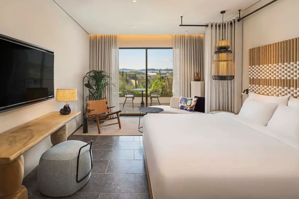 Geräumiges Hotelzimmer mit schlichtem Doppelbett, Sitzecke und Terrasse mit Meeresblick.