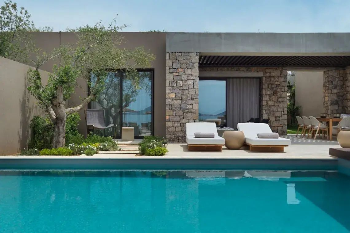 Privater Pool mit zwei Sonnenliegen vor einer modernen Villa mit Steinmauer und Olivenbaum.