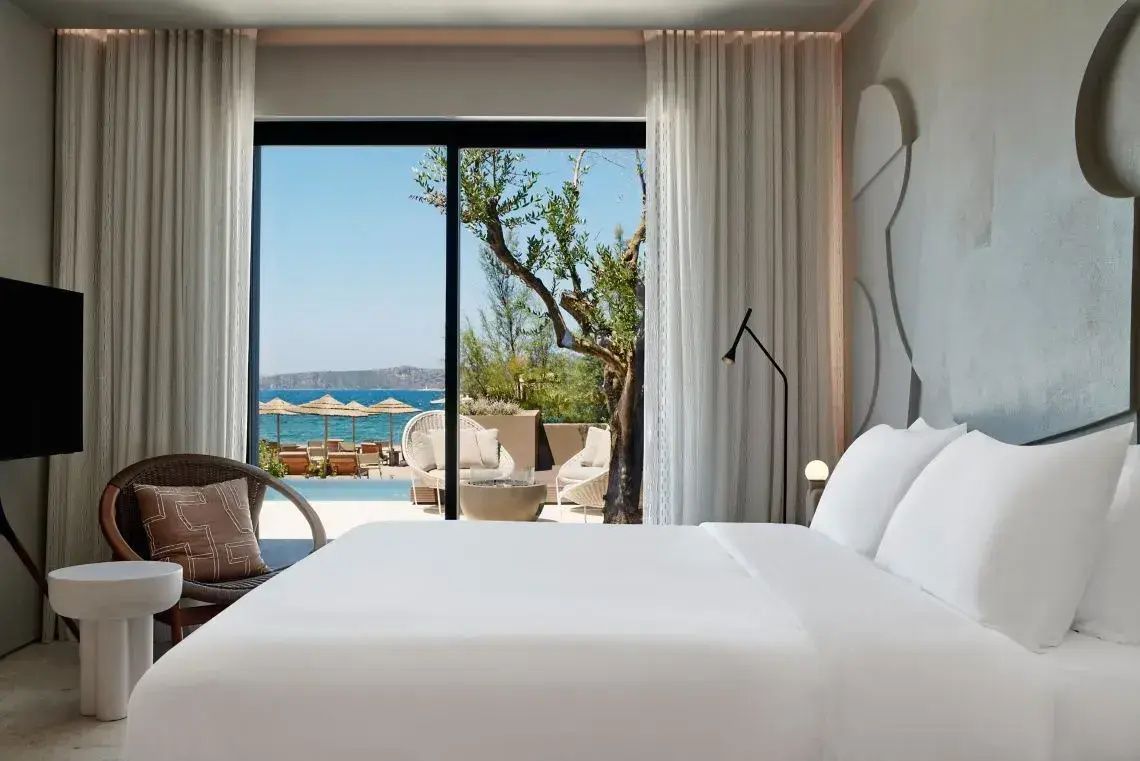 Blick vom Bett durch eine Glasschiebetür auf eine Terrasse mit Pool und das Meer.