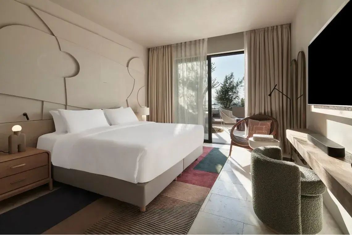 Helles Hotelzimmer mit weiß bezogenem Doppelbett, Sessel und Blick auf die Terrasse.